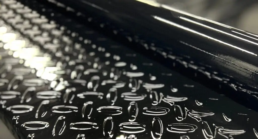 Close-up of the gloss black finish on a Texline trailer's diamond plate deck and frame rail, reflecting the shop lights.