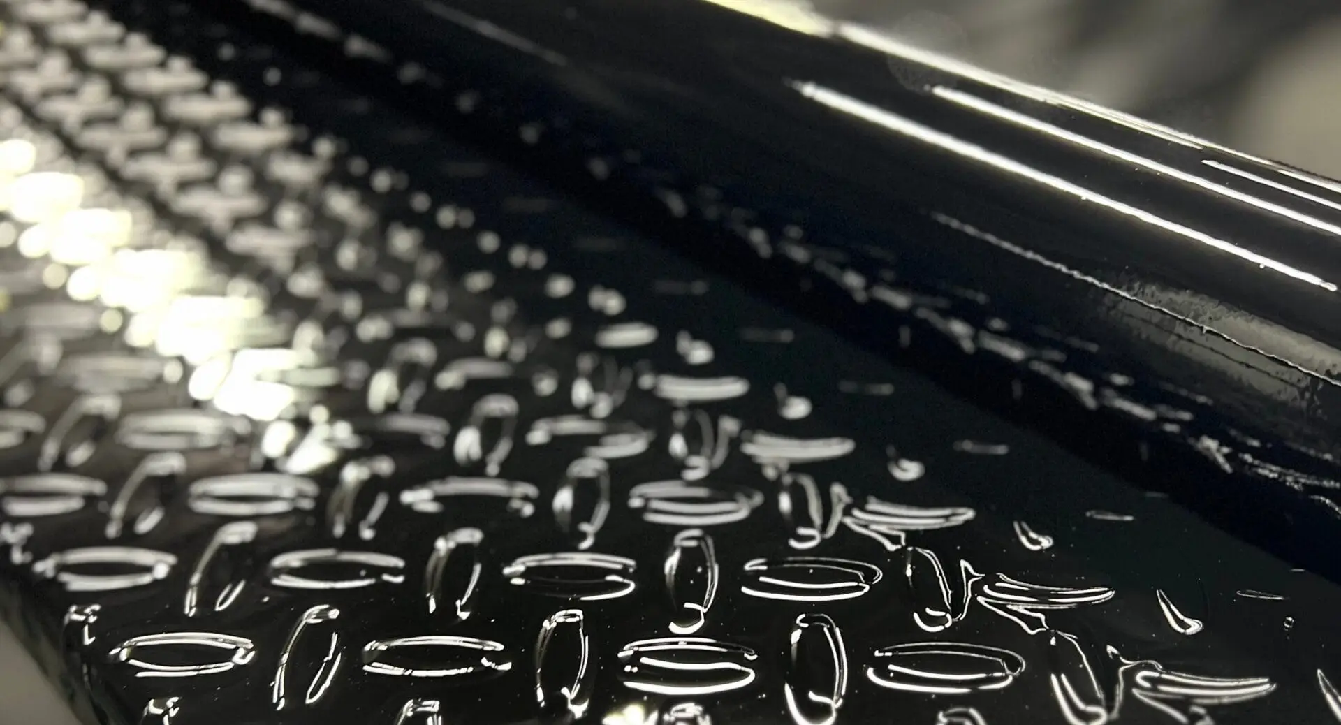 Close-up of the gloss black finish on a Texline trailer's diamond plate deck and frame rail, reflecting the shop lights.